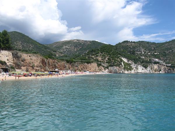 Crowded beach beneath green hills, turquoise sea with white clouds