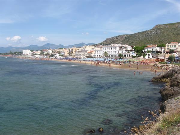 Beachgoers on sandy shore in front of hotels, rocks and turquoise water foreground