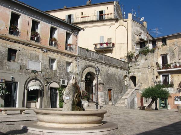 Stone fountain on square, colorful houses with arches and staircase, sunny light
