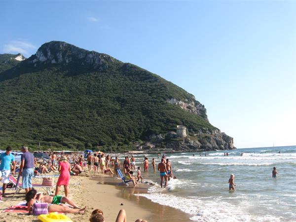 Bathers on sand and in waves under a green mountain with a tower