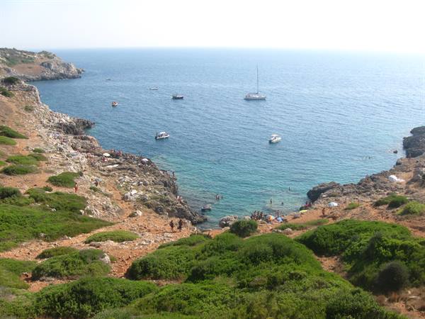 Boats anchored in a rocky cove with cliffs, Mediterranean shrubs and swimmers