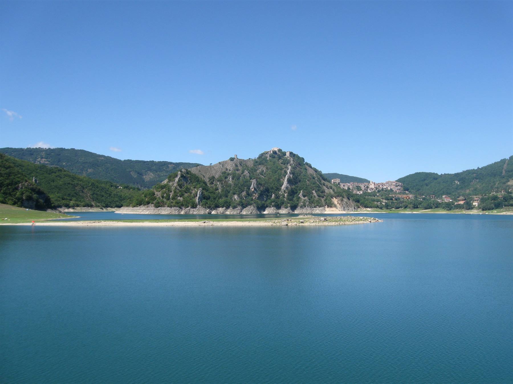 Lago turchese circondato da colline verdi e rocciose sotto cielo azzurro