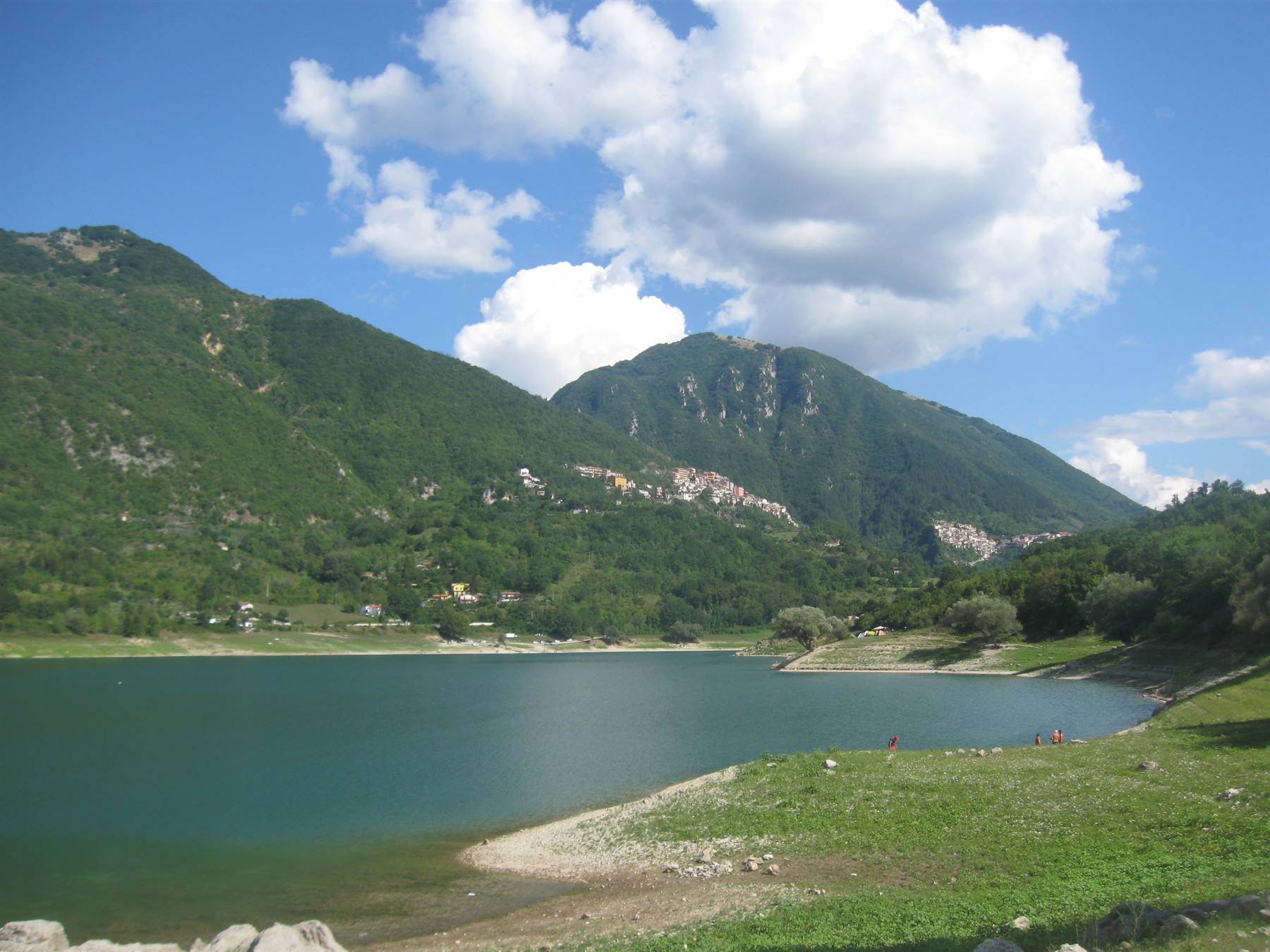 Lago Scanno con verde sponda erbosa, monti boscosi e paese arroccato sotto nuvole