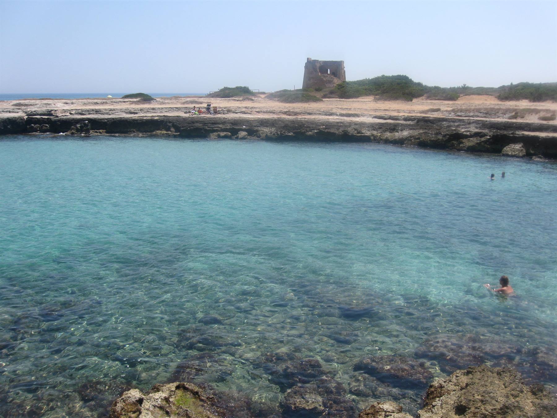 Nuotatore in acqua turchese tra scogli, torre costiera antica sulla riva