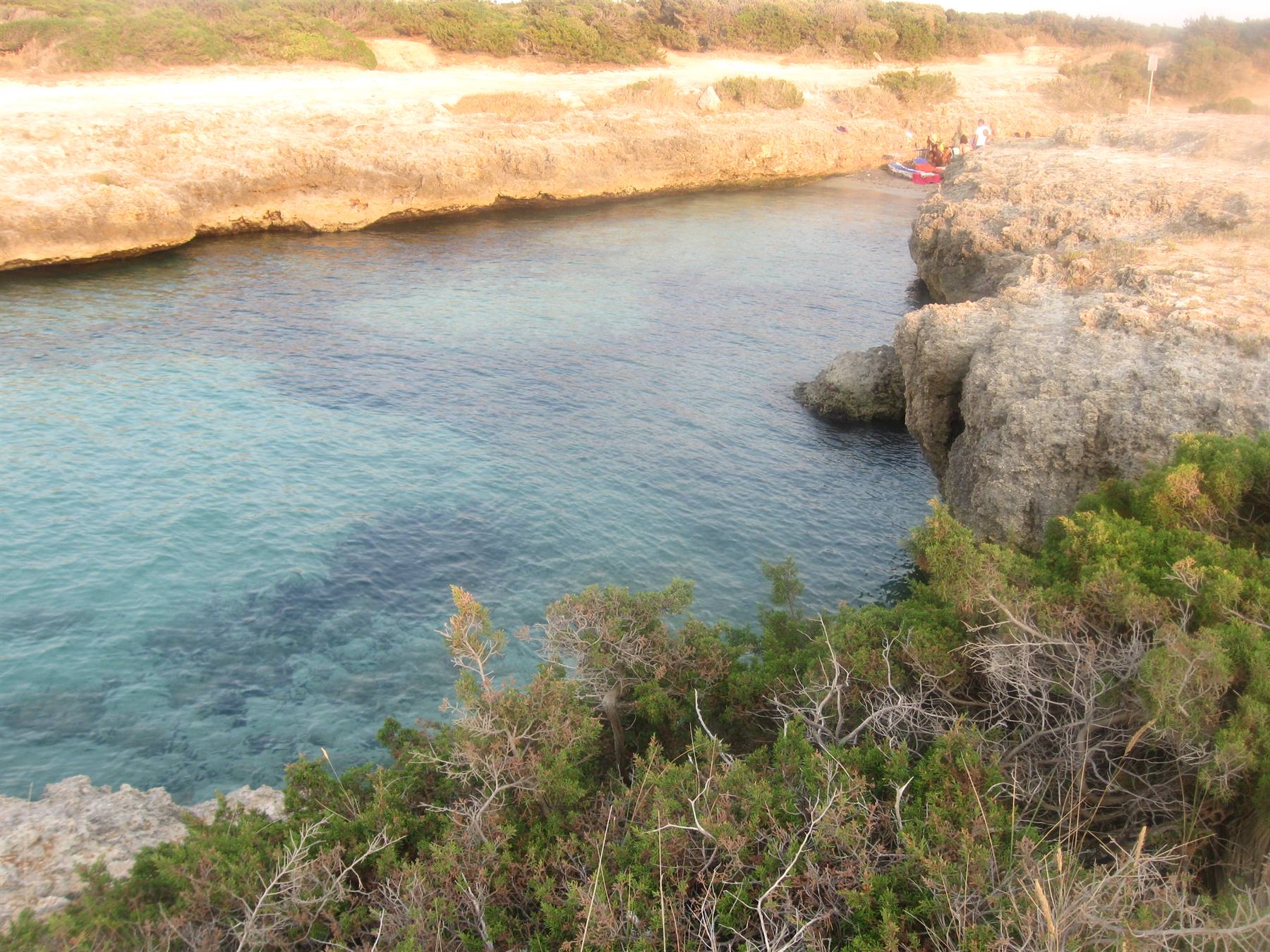 Acqua turchese tra scogli calcarei, macchia mediterranea e barche sotto luce calda