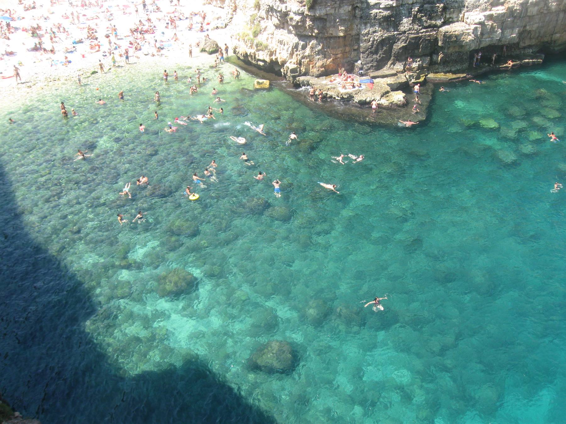 Bagnanti in acqua turchese trasparente sotto falesia rocciosa e spiaggia affollata