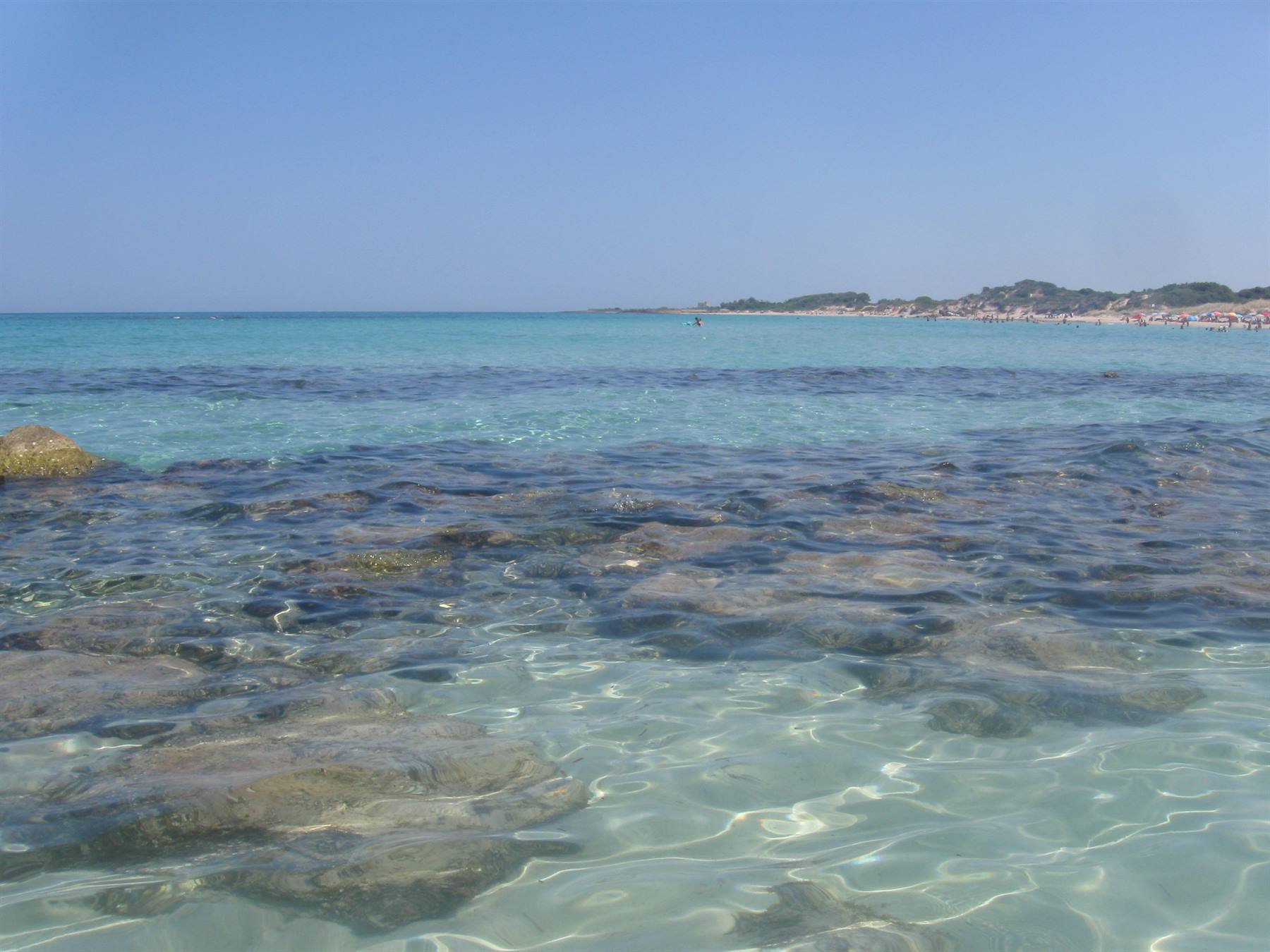 Acqua turchese trasparente su scogli, spiaggia affollata sulla costa sarda