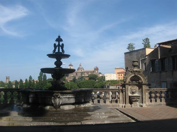 Stone fountain with lion and tiered basins, churches and rooftops behind