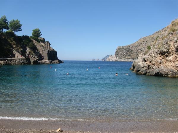 Swimmers in turquoise water between rocky cliffs, Capri faraglioni in the distance