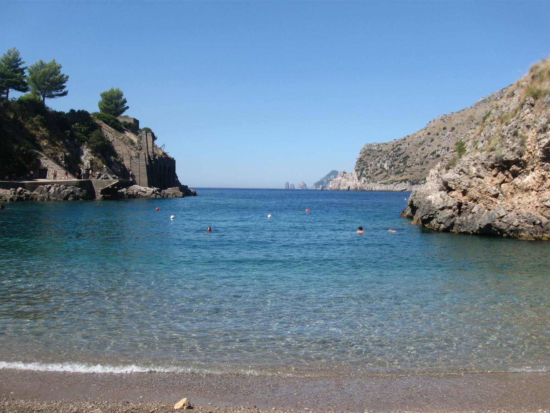 Bagnanti nell'acqua turchese tra scogliere, al largo i faraglioni di Capri
