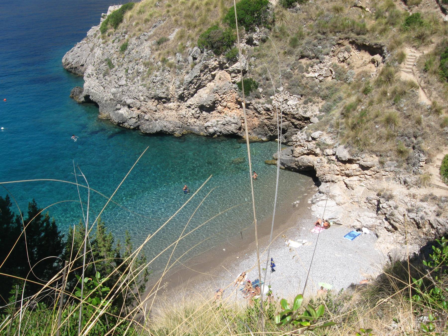 baia con spiaggetta di sassi, bagnanti in acqua turchese tra scogli erbosi