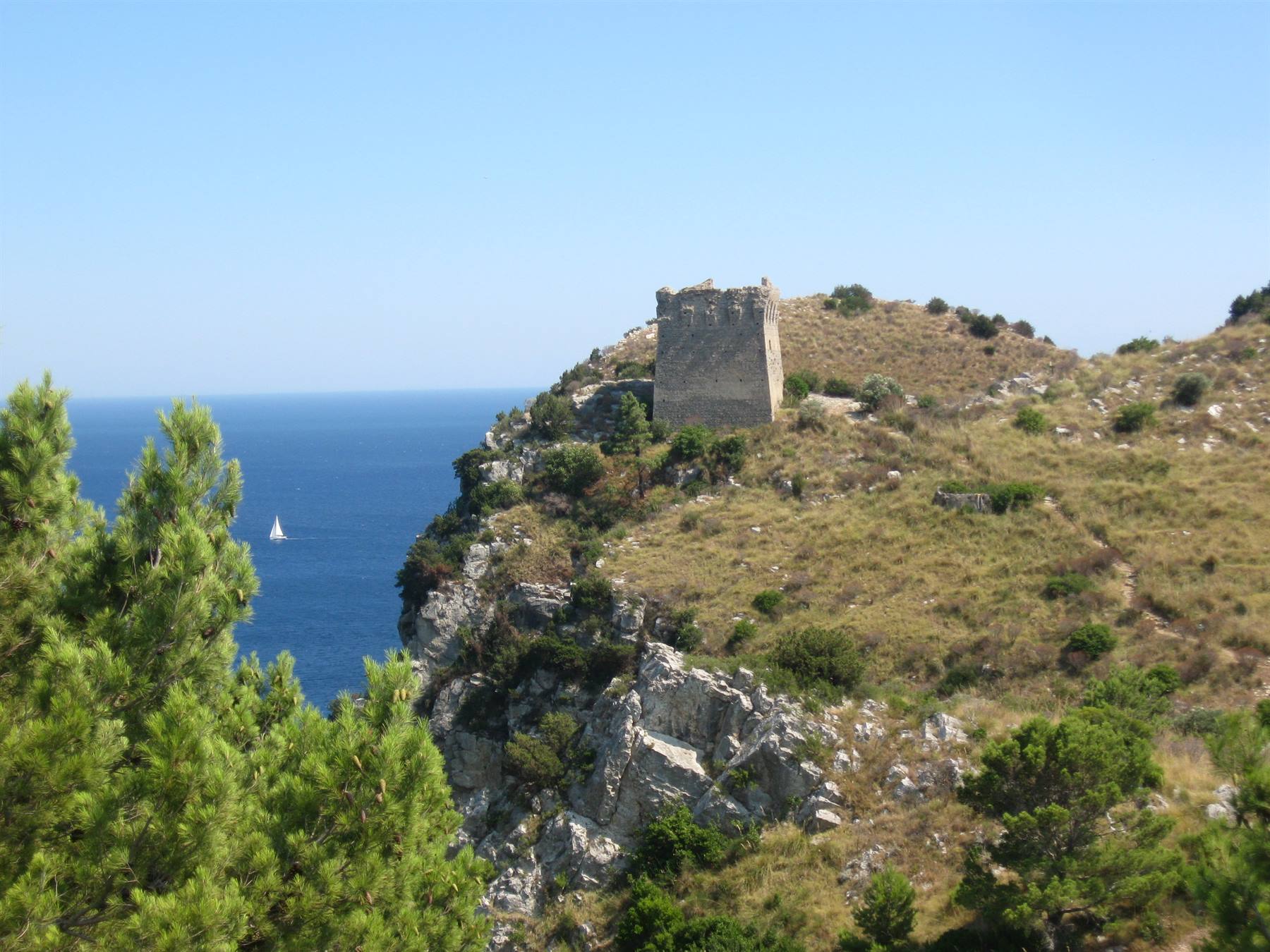 Antica torre di pietra su scogliera a strapiombo sul mare, veliero in lontananza