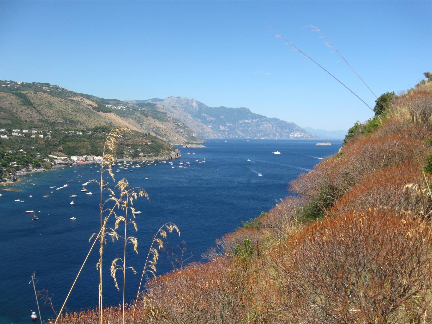 Barche ancorate in una baia blu tra colline e macchia mediterranea secca