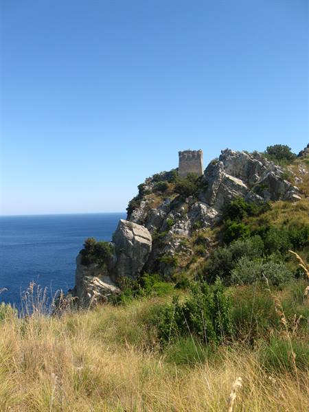 Old stone tower on rocky cliff above blue sea, dry grass under clear sky