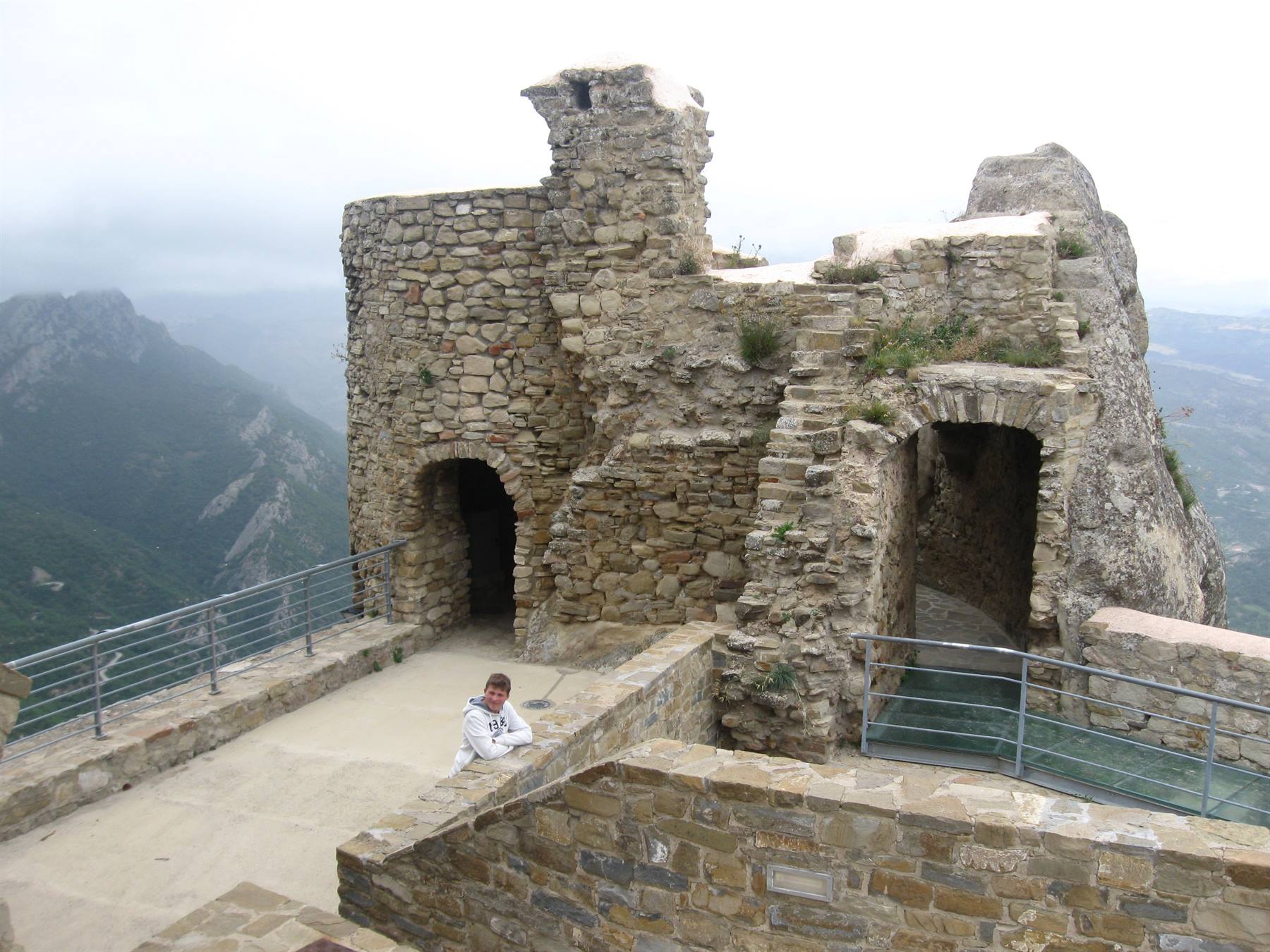 Donna appoggiata al muretto di pietra di una fortezza antica, monti nella nebbia