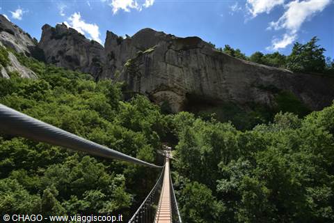 Steel suspension bridge over green forest, rocky cliffs under sunny sky