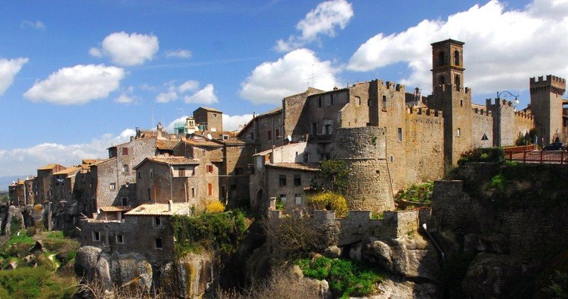 Borgo medievale in pietra su parete di tufo, torri e campanile sotto nuvole bianche, Vitorchiano