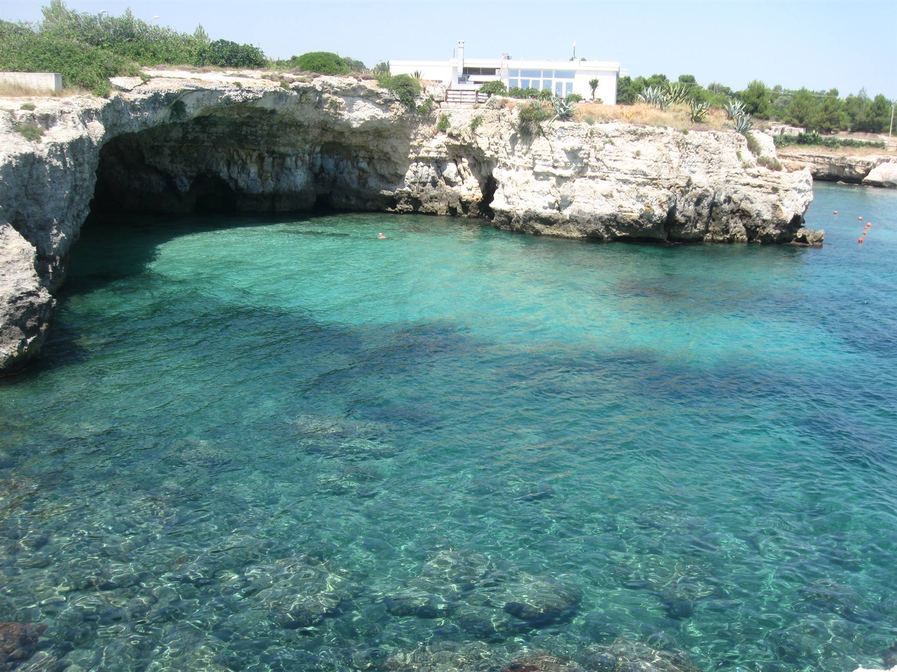 Cala di roccia calcarea con grotta, acqua turchese trasparente, villa sopra la scogliera