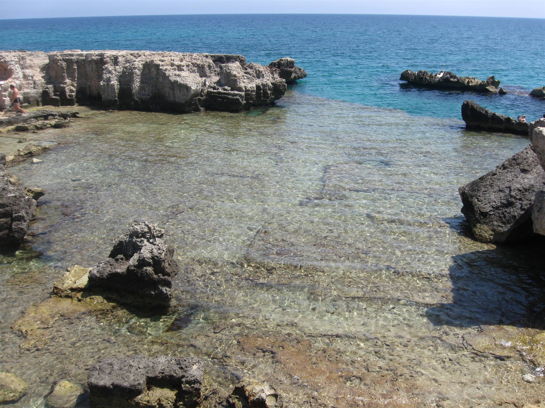 Vasca di roccia calcarea con acqua cristallina, scogli grigi e mare azzurro