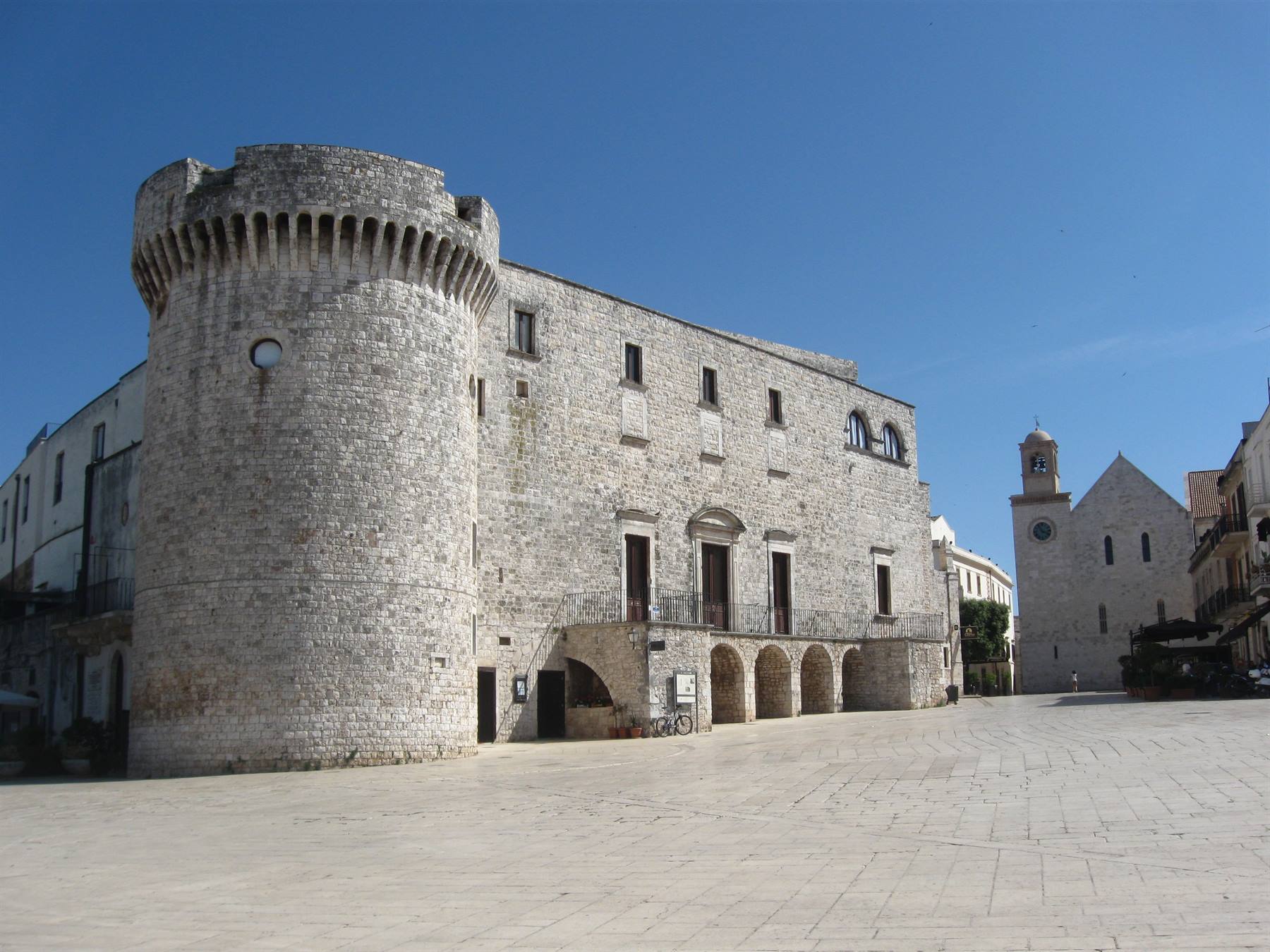 Castello di Conversano, torre circolare in pietra chiara su piazza vuota sotto cielo azzurro