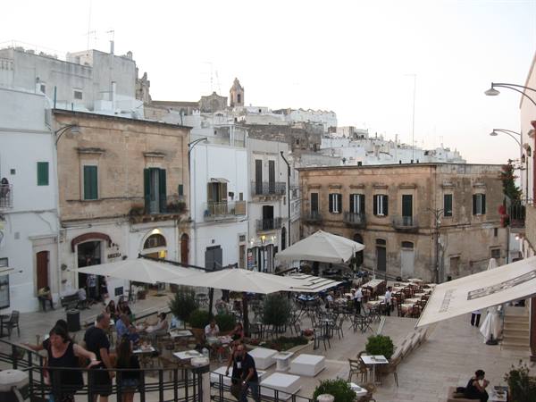 Square with umbrella-shaded café tables among whitewashed houses in Ostuni, bell tower above