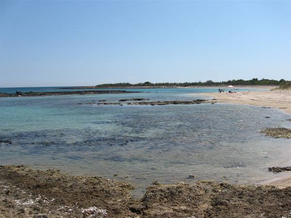 Clear turquoise water over rocks, sandy beach with swimmers, blue sky