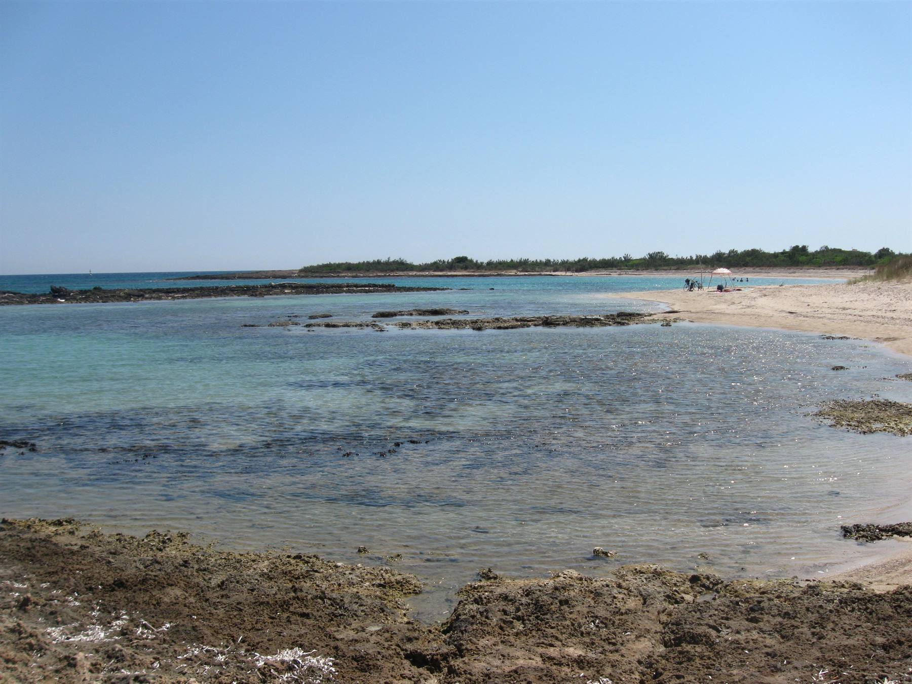 Acque turchesi trasparenti su scogliera, spiaggia sabbiosa con bagnanti, cielo azzurro