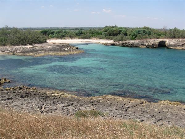 Turquoise water between limestone rocks and Mediterranean scrub, sandy beach behind