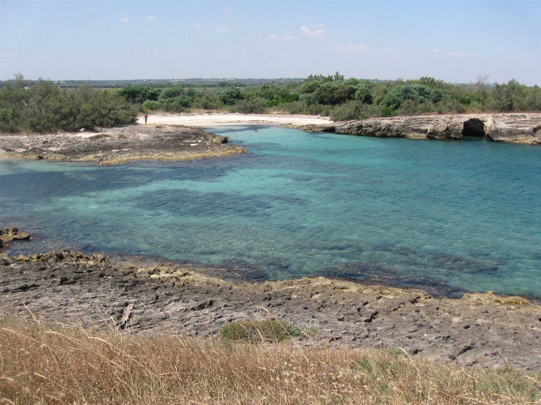Acqua turchese tra scogli calcarei e macchia mediterranea, spiaggia sabbiosa al fondo
