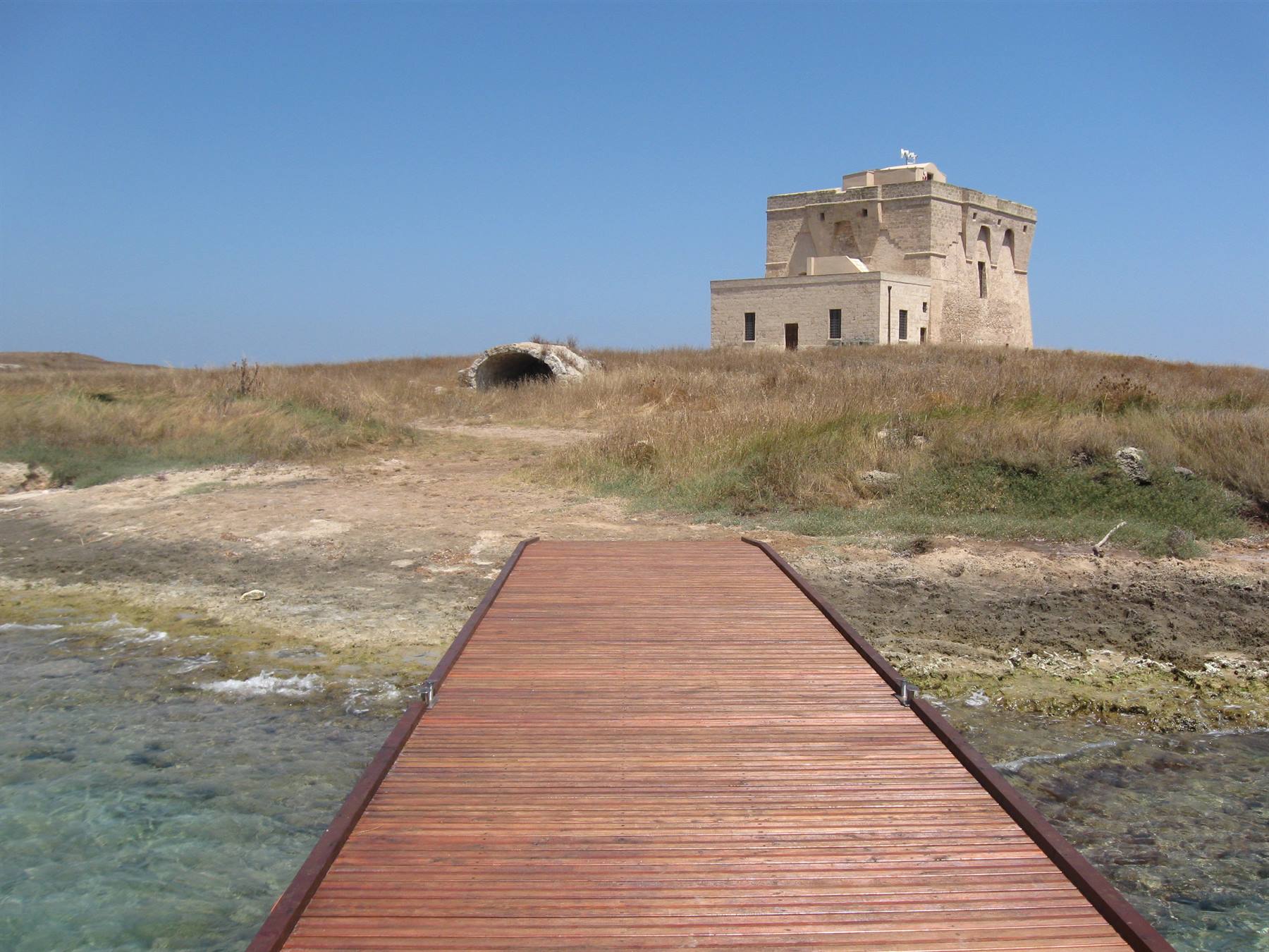 Passerella di legno verso antica torre costiera in pietra sotto cielo azzurro