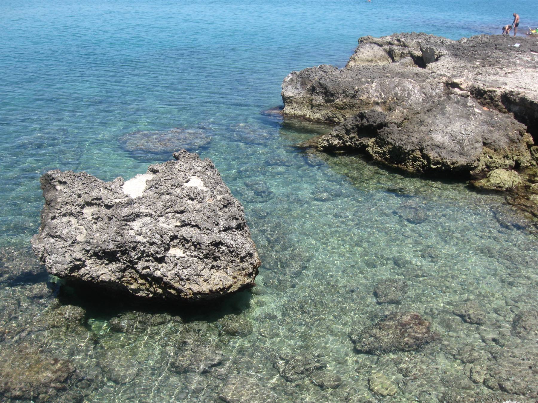 Scogli neri nel mare trasparente, bagnanti in fondo a destra