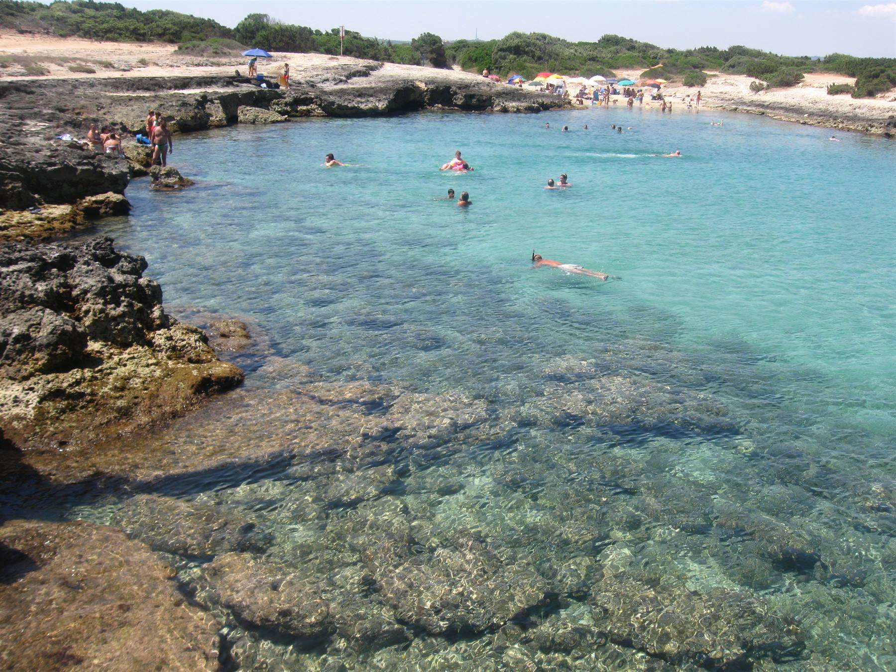 Bagnanti in acqua turchese trasparente tra scogli calcarei e sabbia bianca, Puglia