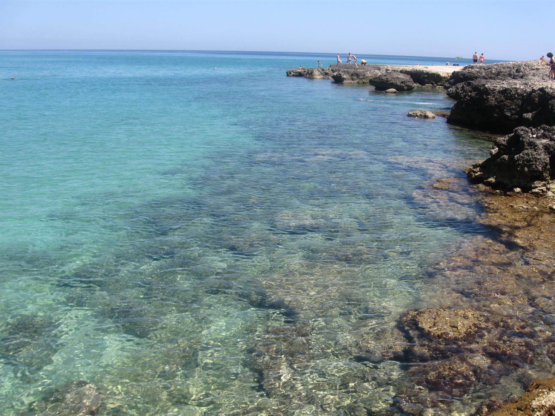 Acqua turchese trasparente su scogli, bagnanti sulla scogliera a destra