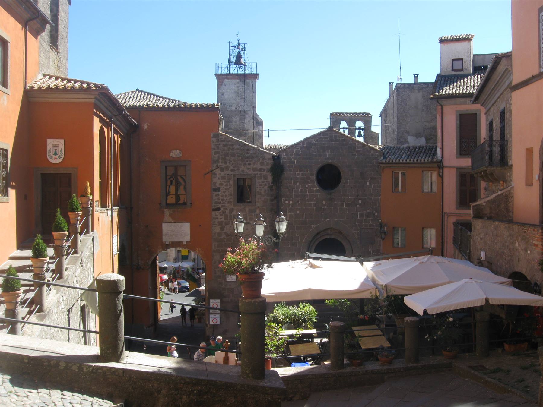 Piazzetta medievale in pietra con chiesa, campanile e ombrelloni bianchi dei caffè