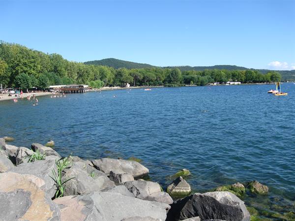Dark boulders foreground, blue lake with swimmers and pedal boats on the beach