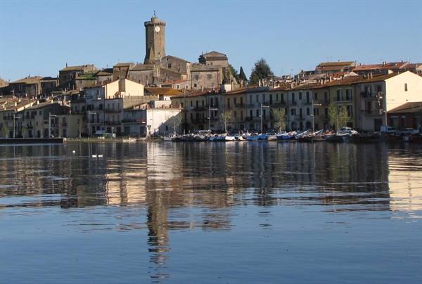 Boats moored in front of stone houses with medieval tower, reflected on calm lake