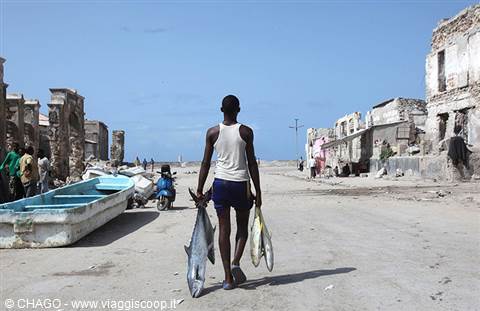 Fisherman carrying freshly caught fish walks past ruined buildings and boats