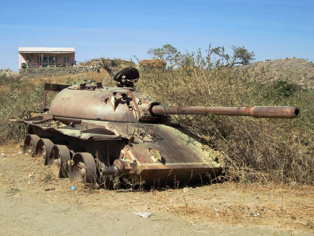 Carro armato arrugginito abbandonato tra cespugli secchi, casa bianca sullo sfondo