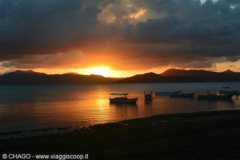Boats floating offshore under dark clouds, sun setting behind mountains reflects on the water