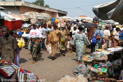 Crowded open-air market, women in colorful dresses carry sacks on their heads past produce stalls