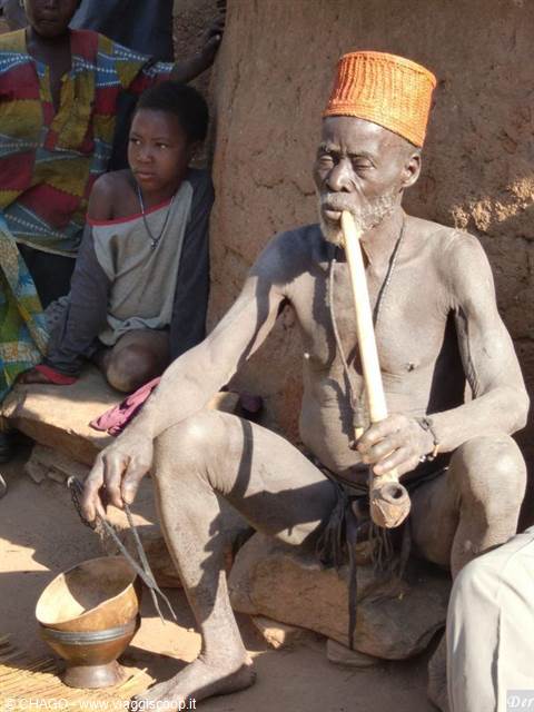 Elderly man with orange woven hat smokes a long wooden pipe beside a child, in front of mud houses