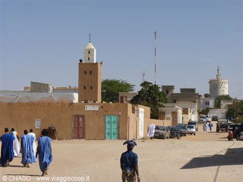 Men in blue boubous walk toward an earthen mosque with white minaret