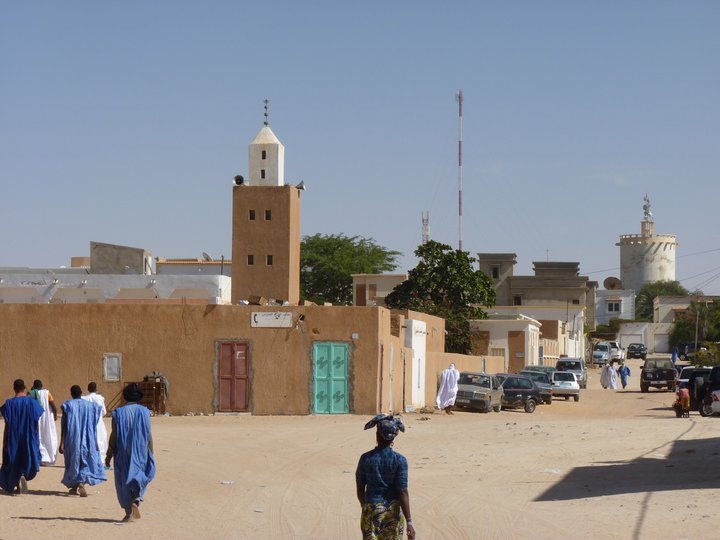 Uomini in boubou blu camminano verso una moschea di terra con minareto bianco