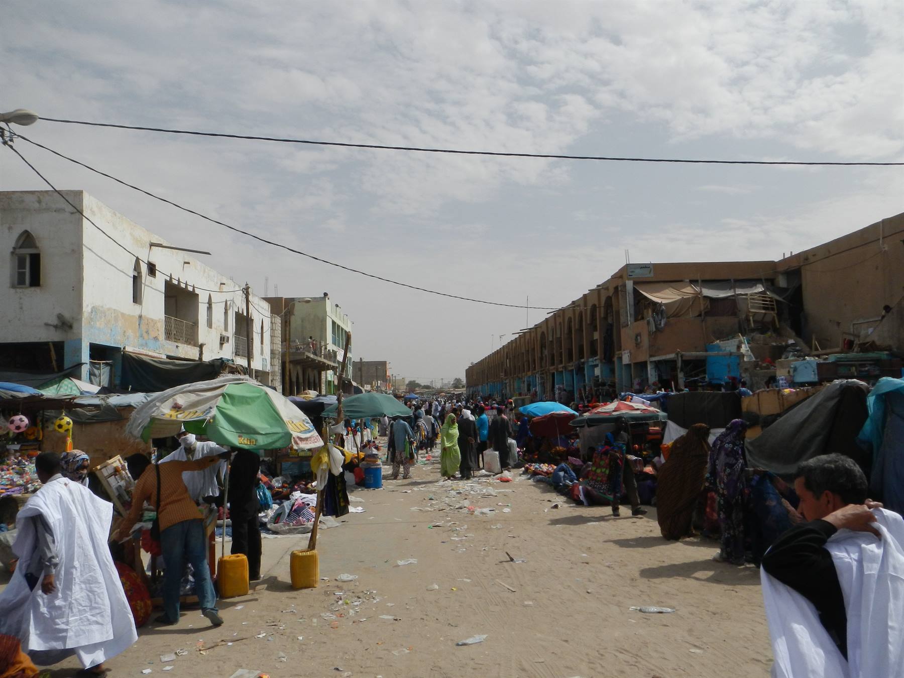 Mercato di strada sabbioso, ombrelloni verdi, bancarelle, edifici bassi, gente in vestiti bianchi