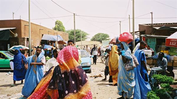 Women in colorful boubous at a street market, brick buildings, harsh light