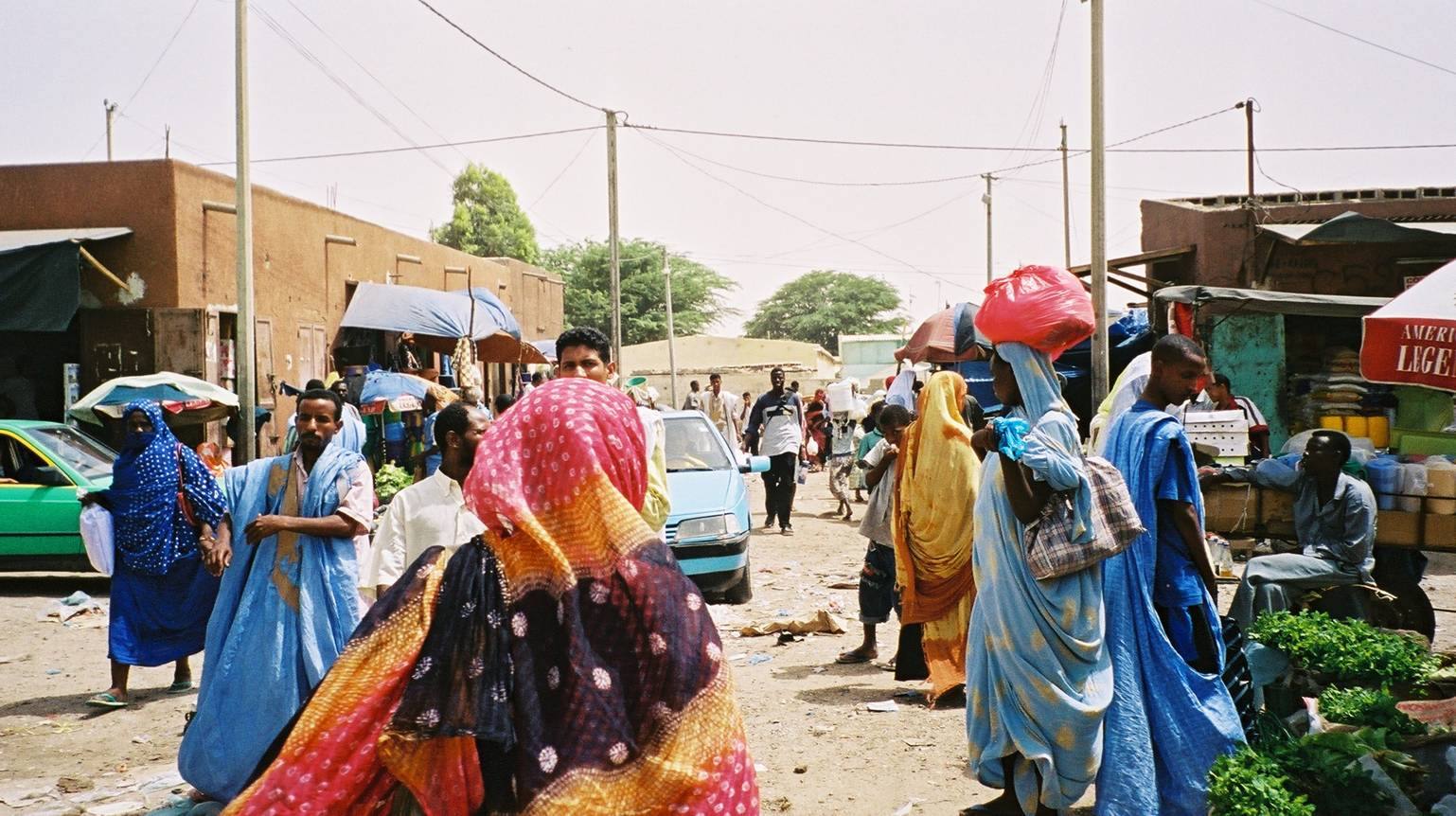 Donne in boubou colorato al mercato di strada, case in mattone, luce intensa