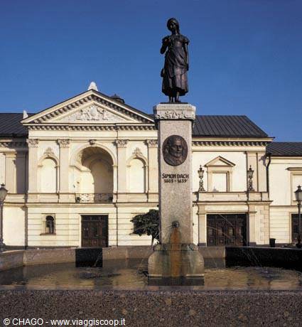 Bronze statue on stone pedestal before a white palace, sunny fountain