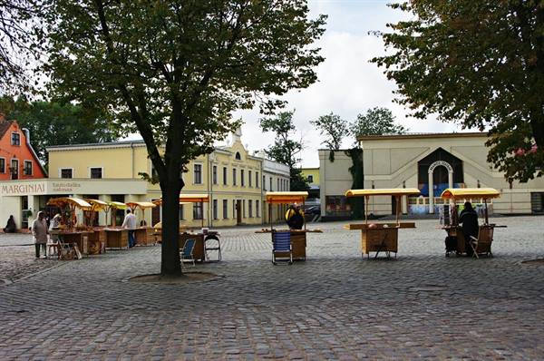 Cobblestone square with wooden market kiosks under trees, yellow buildings and cloudy sky