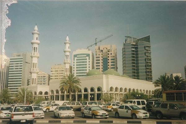 White mosque with minarets and green domes, skyscrapers and taxis in front