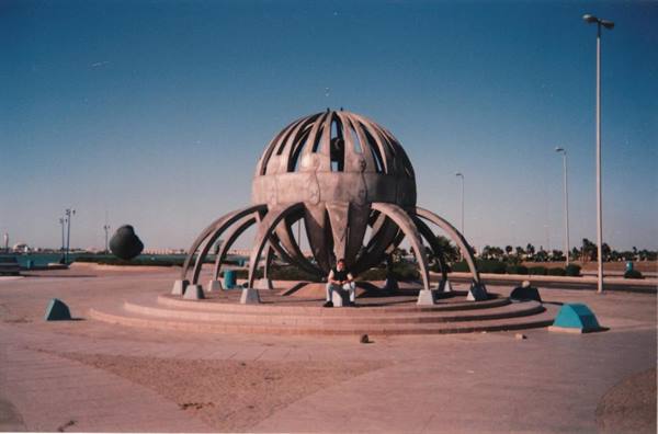 Man sitting under a large metal globe-shaped sculpture on the waterfront